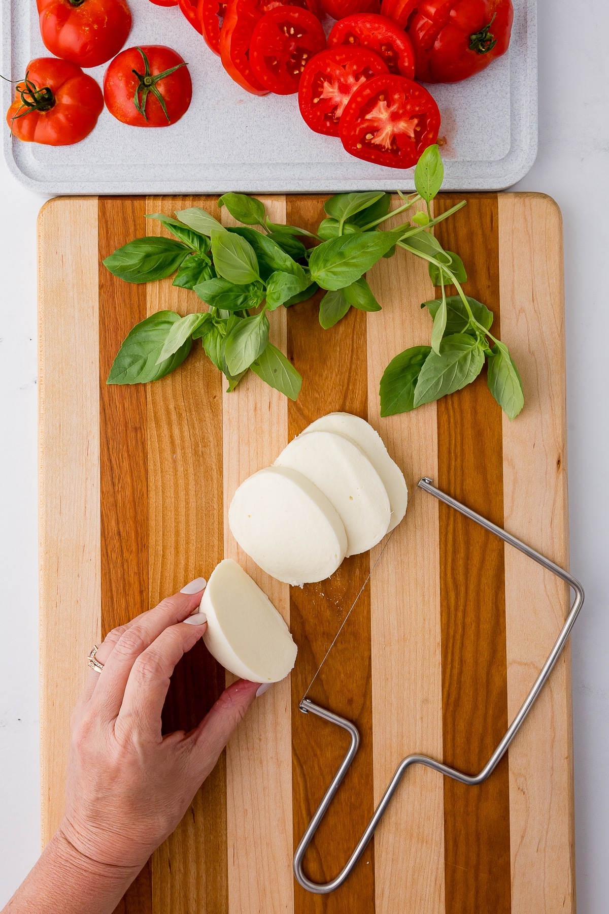 A hand slicing mozzarella cheese on a wooden cutting board with basil and tomatoes