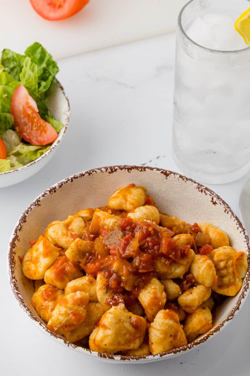 bowl of ñochis in a pasta bowl with tomato pasta sauce, a side salad, and a glass of ice water with a lemon