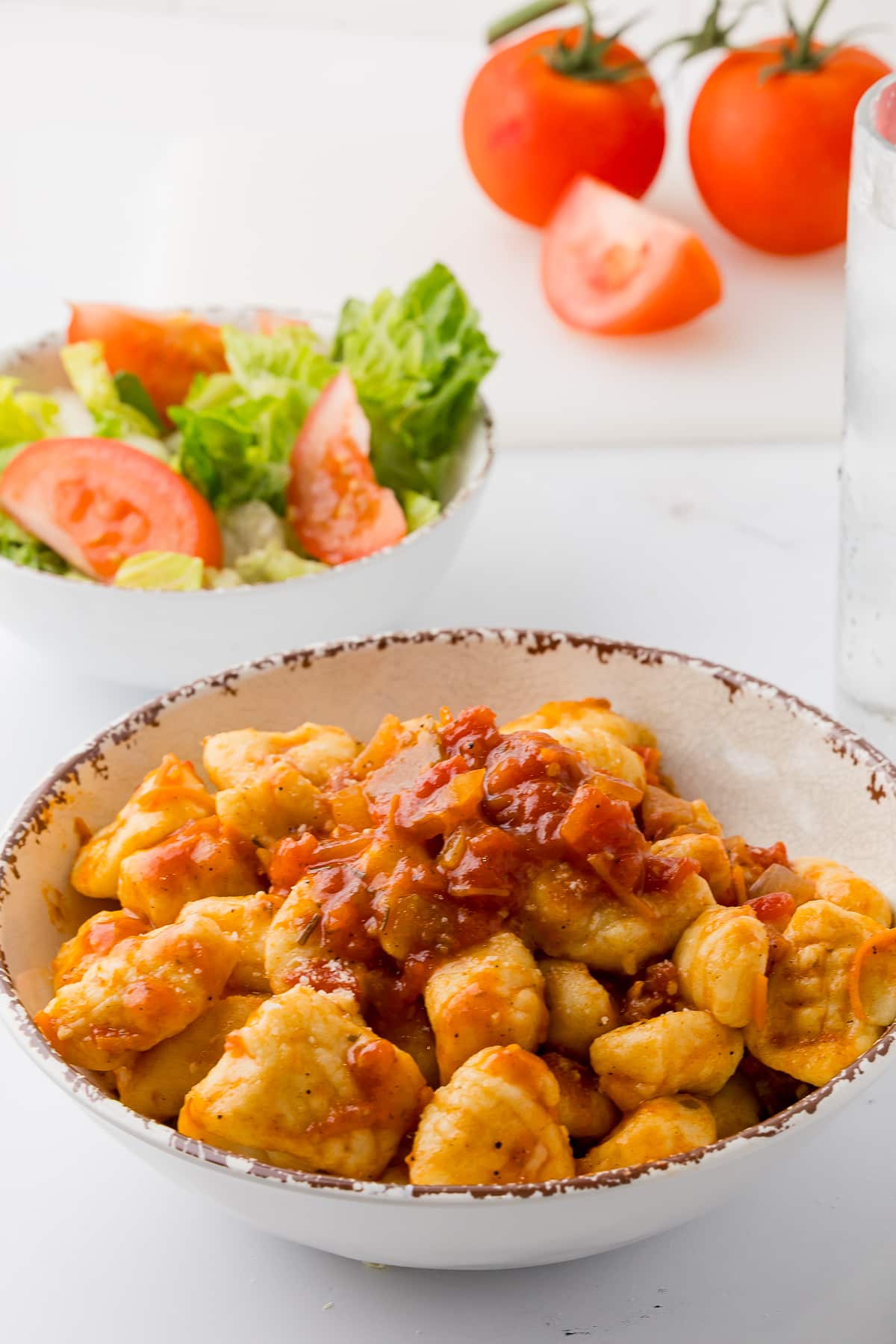 homemade gnocchis in a white pasta bowl with a side salad and tomatoes in the background