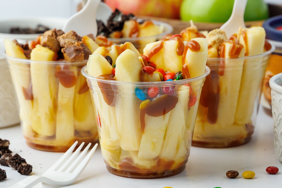 Close up of sliced apples in clear plastic cups topped with caramel and mini m&ms and crushed candy bars