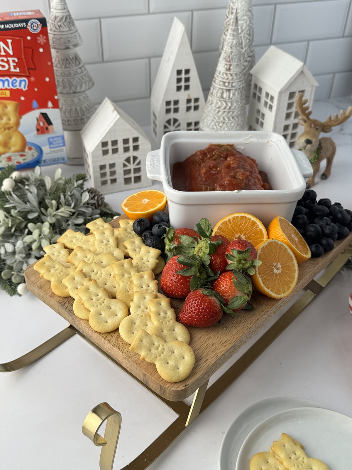 Wooden sled cutting board with snowman crakers, strawberries, oranges, and cream cheese and salsa dip on a white counter with white snow village behind it