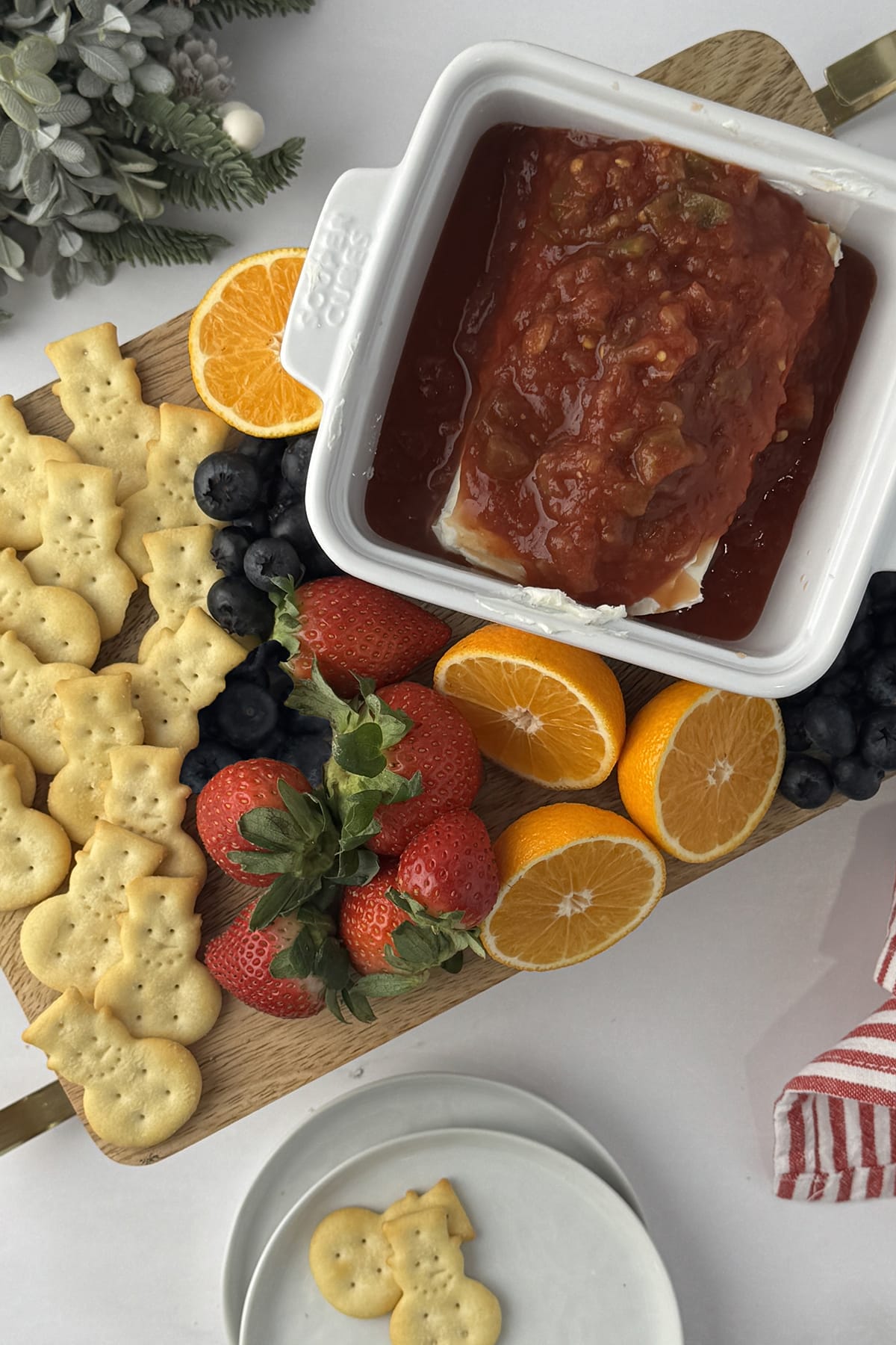 overhead photo of Wooden sled cutting board with snowman crackers, strawberries, oranges, and cream cheese and salsa dip on a white counter