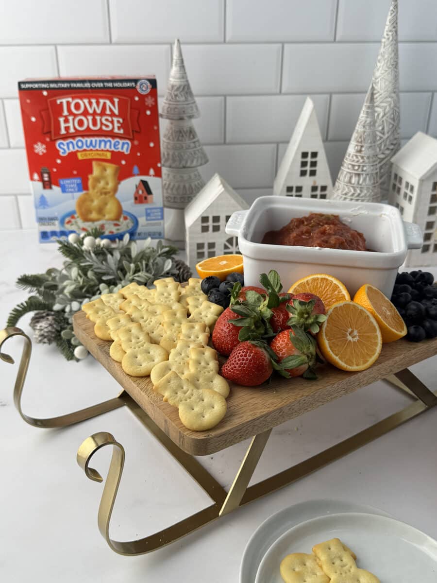 Wooden sled cutting board with snowman crackers, strawberries, oranges, and cream cheese and salsa dip on a white counter