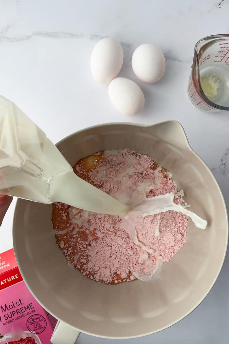 batter bowl with strawberry cake mix and milk pouring into the bowl with 3 eggs on a white countertop