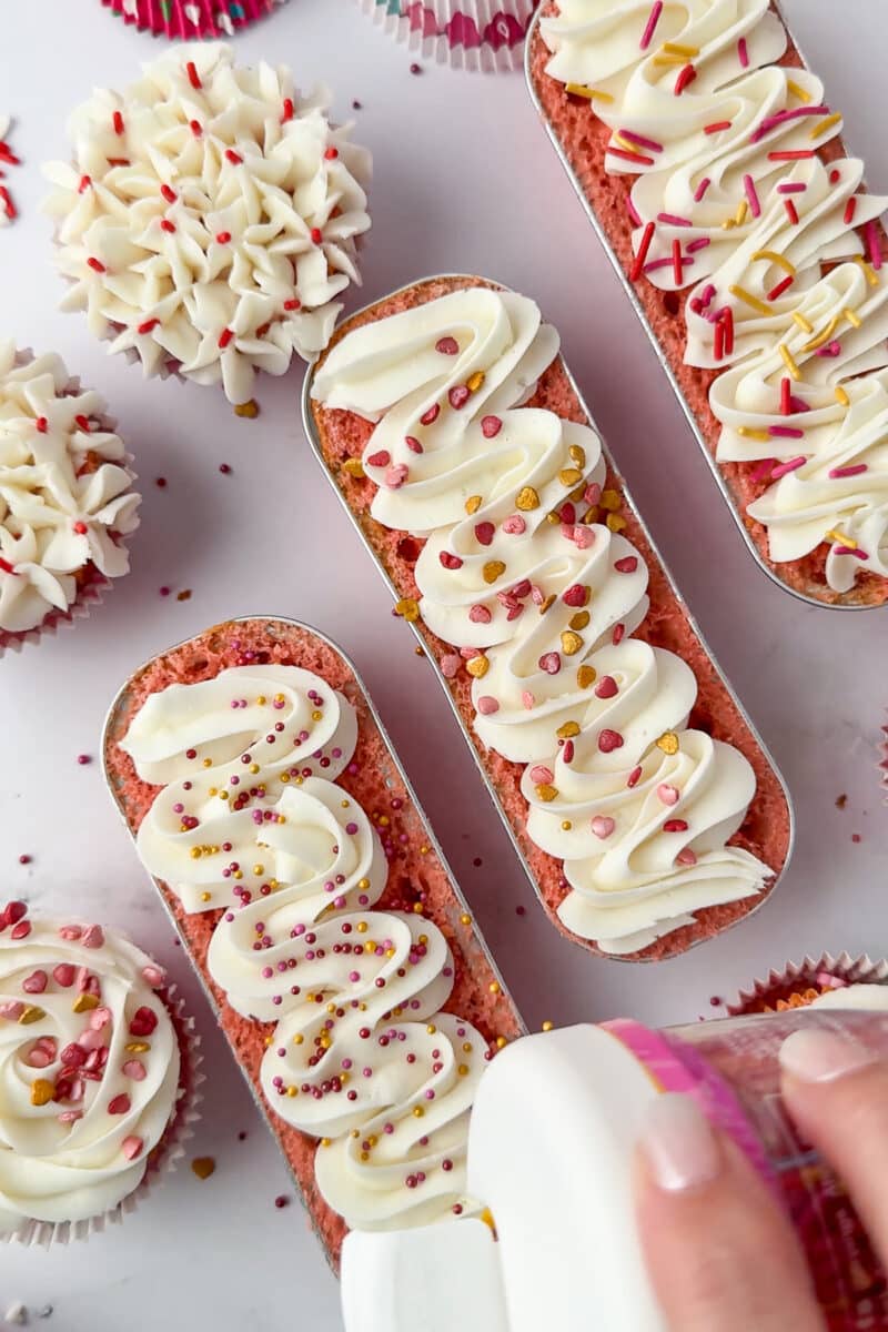 wilton sprinkles being poured onto whiter frosting on a mini loaf pan filled with baked strawberry cake, cupcakes and two other loaves and cupcakes