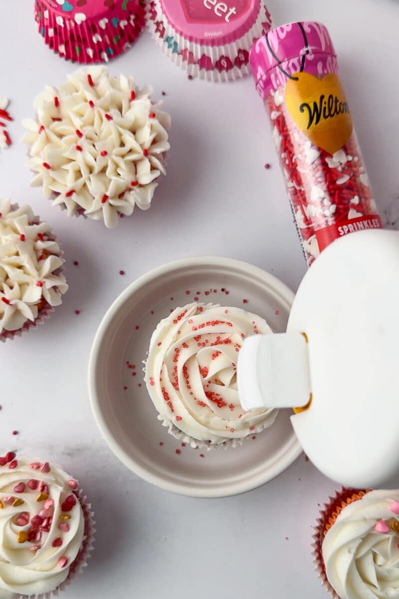 pouring pink sanding sugar over white frosting on a strawberry cupcake with wilton valentine's day sprinkles