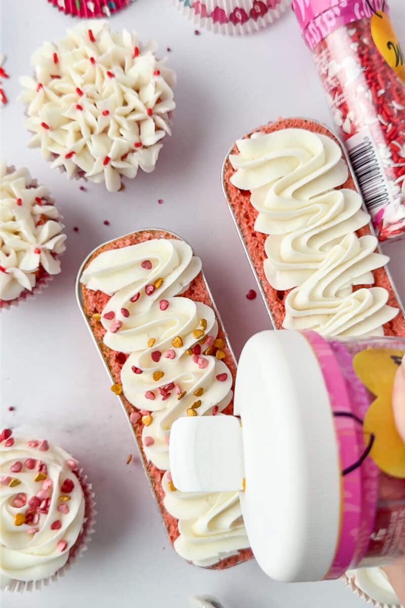 wilton sprinkles being poured onto whiter frosting on a mini loaf pan filled with baked strawberry cake