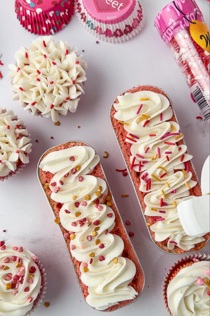 wilton sprinkles being poured onto whiter frosting on a mini loaf pan filled with baked strawberry cake, decorated cupcakes on the countertop
