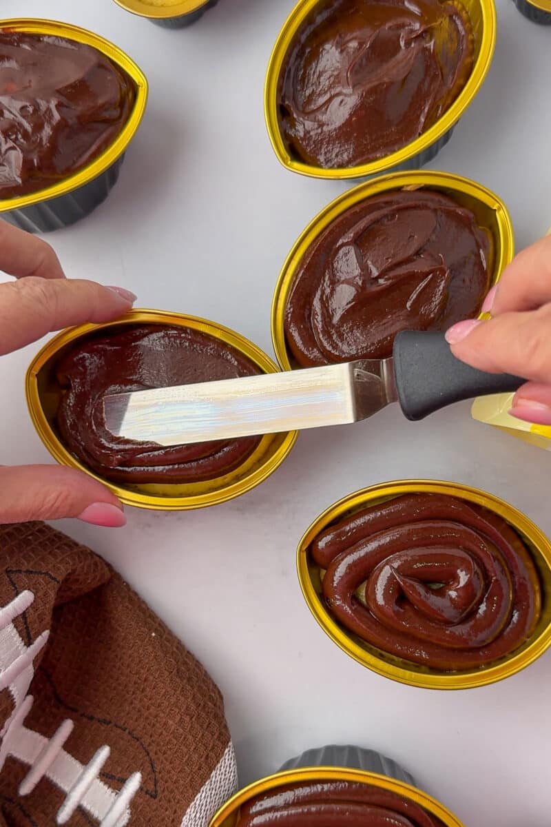 spreading chocolate frosting on a football cupcake with a spatula