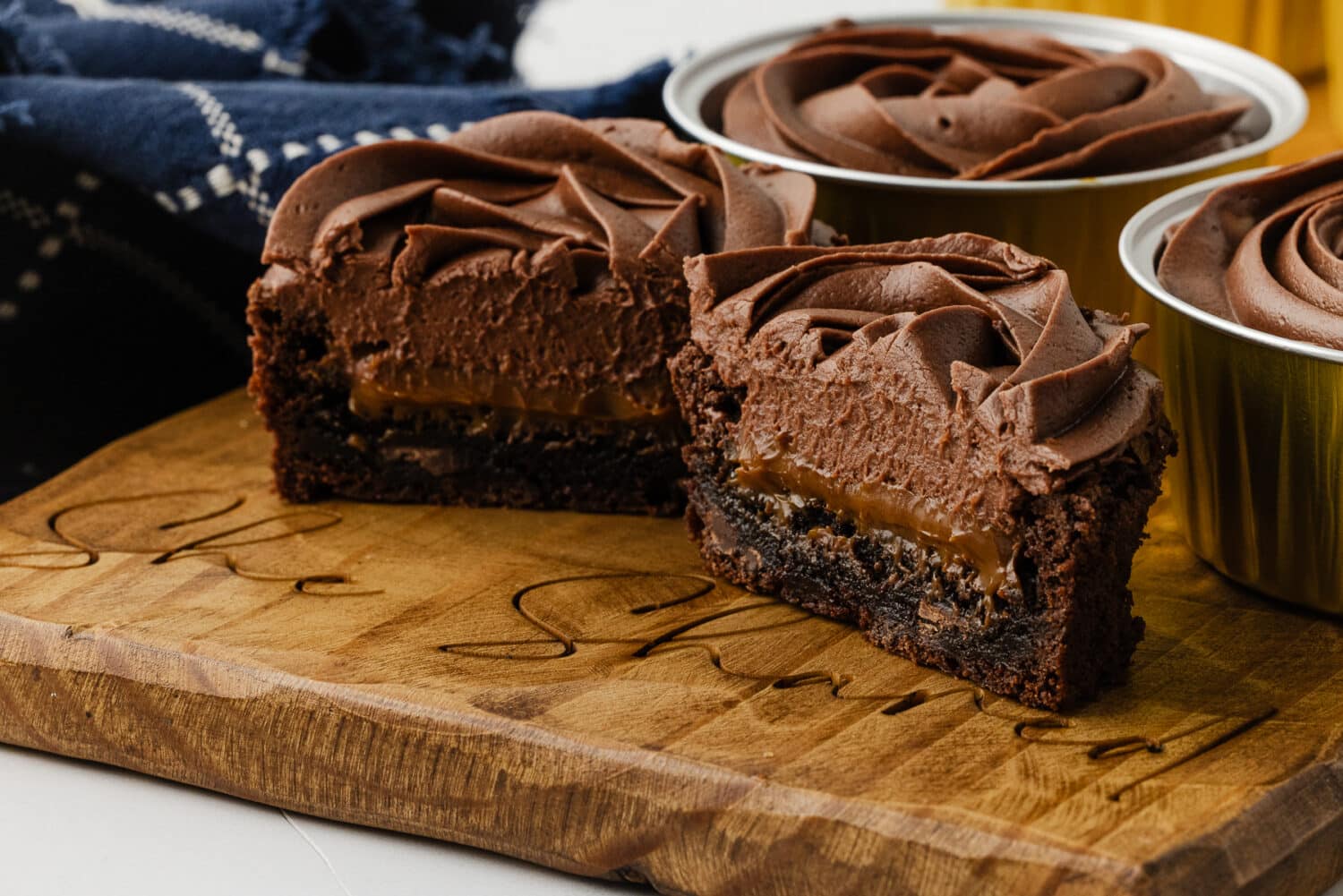 A close-up cross-section of a gluten-free brownie on a "She Shared" engraved wooden board, revealing a thick layer of gooey dulce de leche filling topped with a swirl of chocolate frosting.