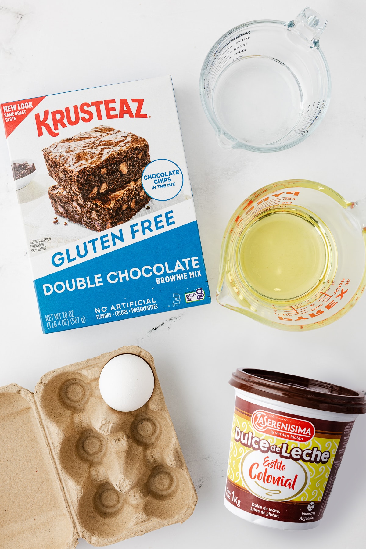 Flat lay of ingredients for gluten-free brownies: Krusteaz Double Chocolate mix, a bowl of oil, a bowl of water, one egg in a carton, and a tub of La Serenisima Dulce de Leche.