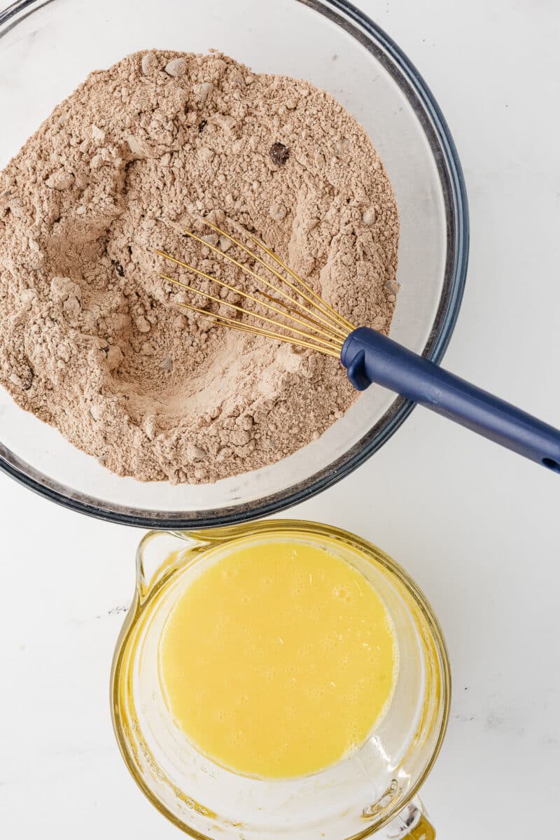 Overhead view of a glass mixing bowl containing dry Krusteaz gluten-free brownie mix next to a smaller glass bowl filled with whisked egg and oil.
