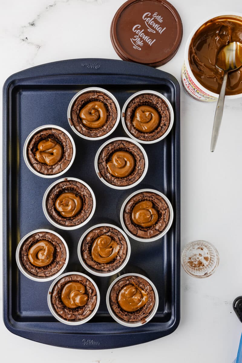 An overhead view of a blue baking sheet with ten brownies in gold foil tins, each with a generous amount of creamy dulce de leche piped into the center wells.