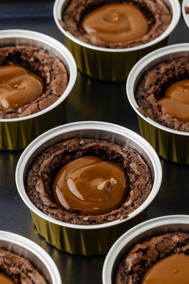 An angled, close-up shot of individual gluten-free brownies in gold tins, highlighting the glossy, smooth texture of the dulce de leche filling.