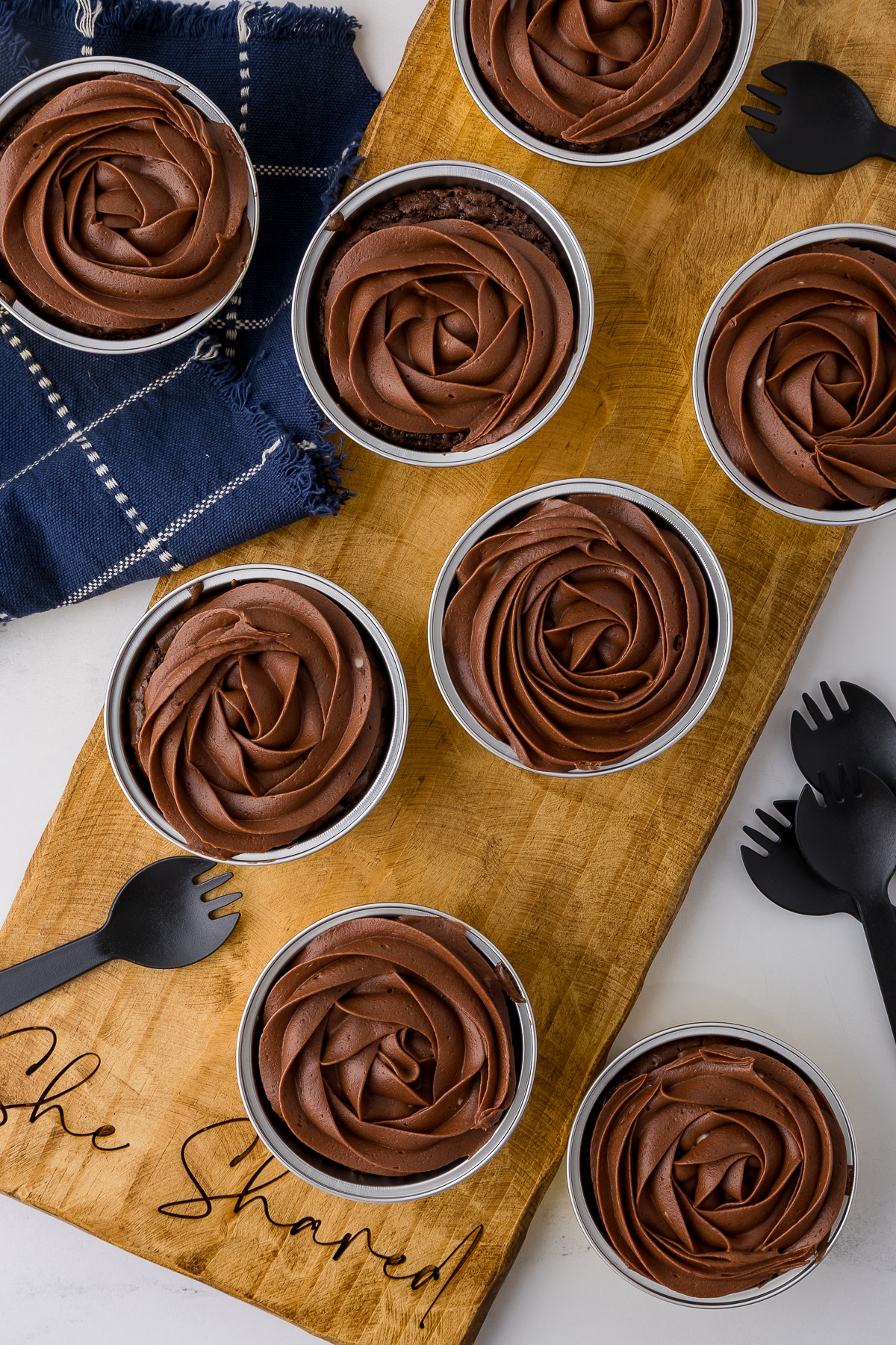 A flat lay of eight gluten-free brownies on a "She Shared" wood board, each topped with a professional rose-shaped swirl of chocolate frosting.