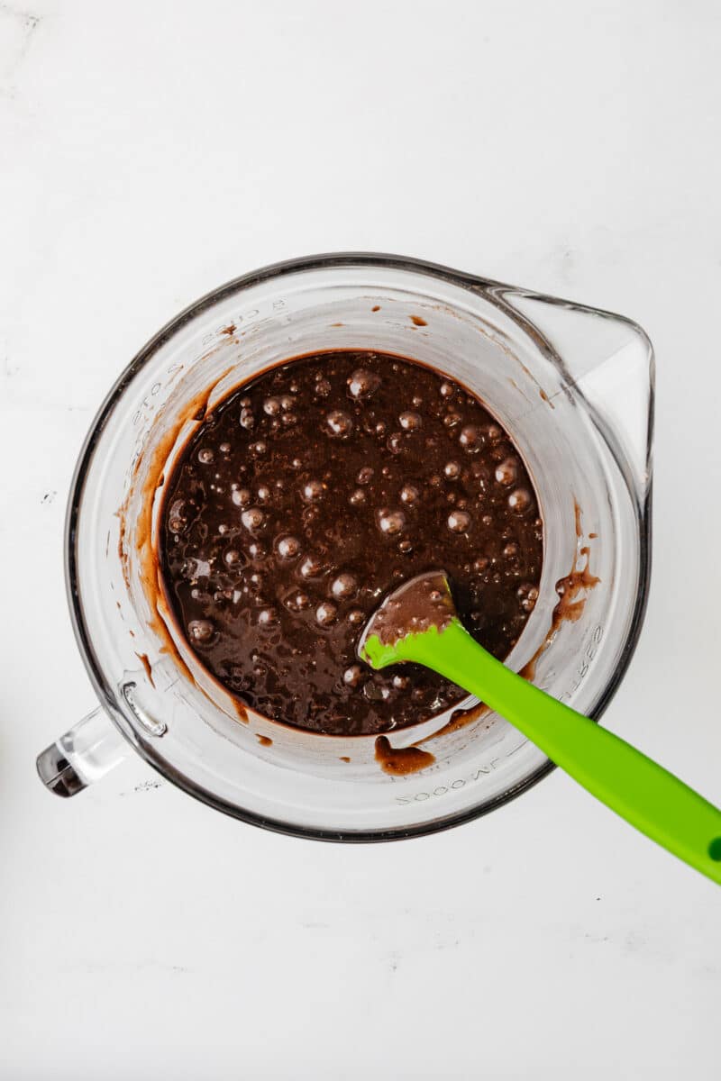 A glass mixing bowl filled with smooth, dark chocolate brownie batter being stirred with a green spatula.