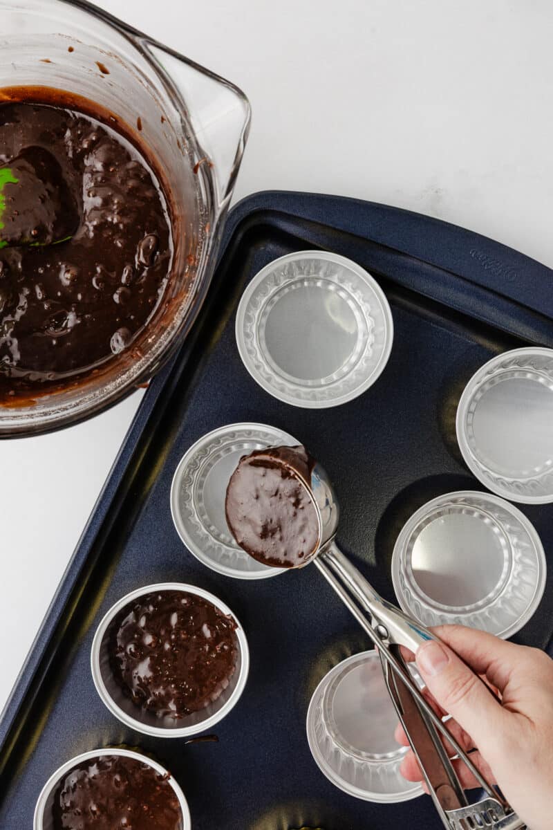 A hand using a metal cookie scoop to portion out dark chocolate brownie batter into individual silver foil baking tins on a dark blue Wilton baking sheet.