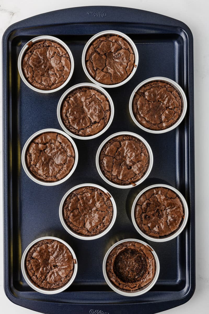 A baking sheet holding ten individual baked brownies in foil tins, with one brownie showing a deep center indentation.