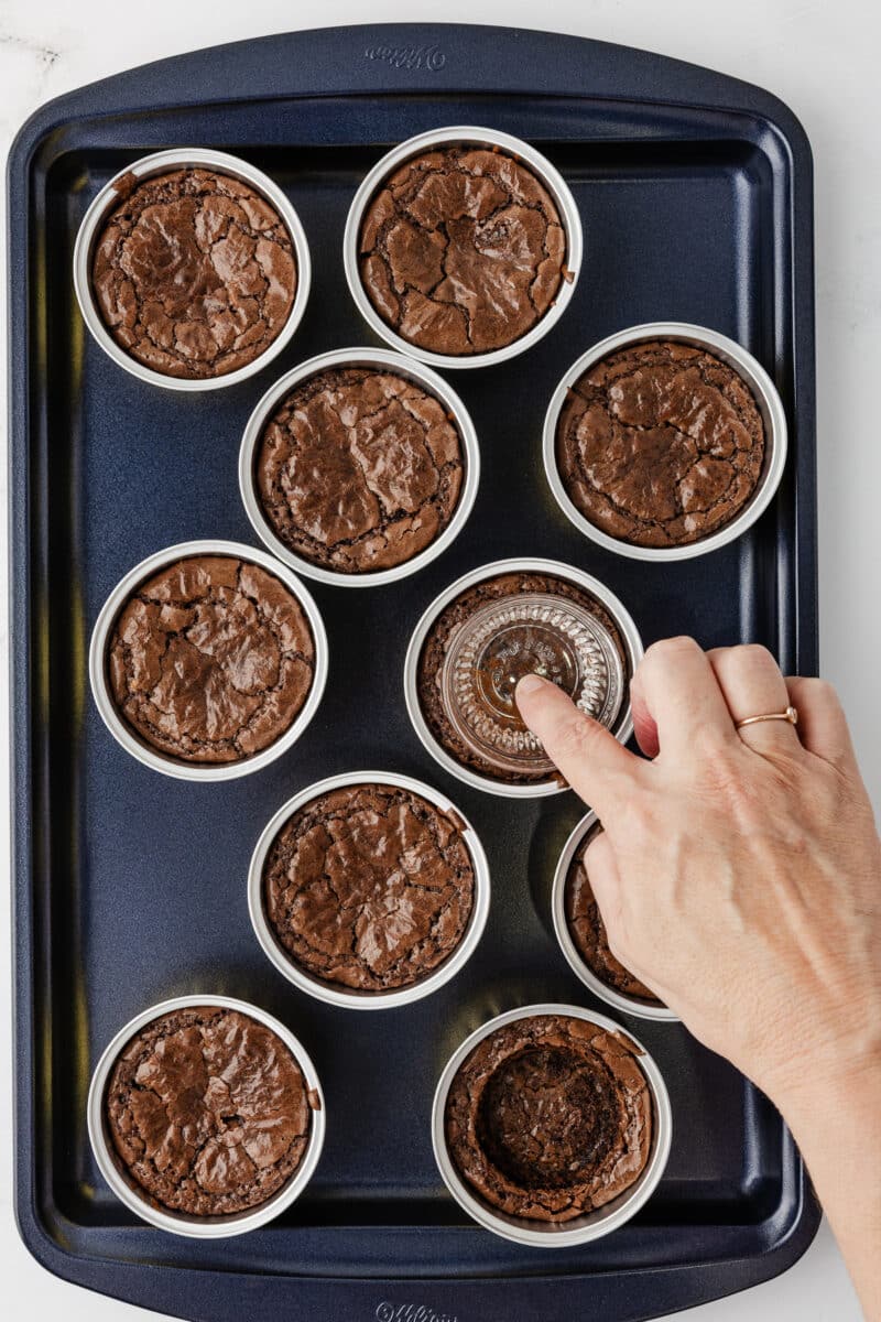 A hand pressing a small glass jar into the center of a warm, individual baked brownie to create a well for the dulce de leche filling.