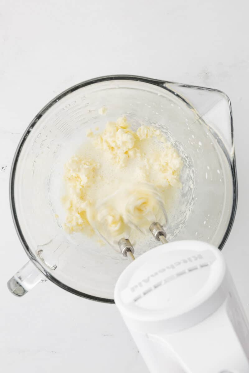 An electric KitchenAid hand mixer whipping softened butter and milk together in a glass bowl until the mixture is creamy and pale.