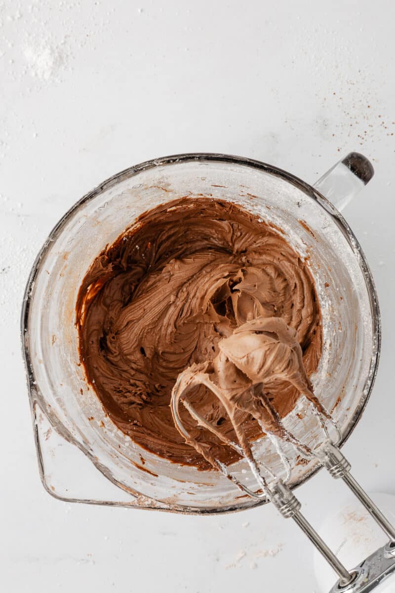 A glass mixing bowl showing the final, creamy chocolate buttercream frosting with the whisks of a hand mixer lifted to show a thick, smooth peak.