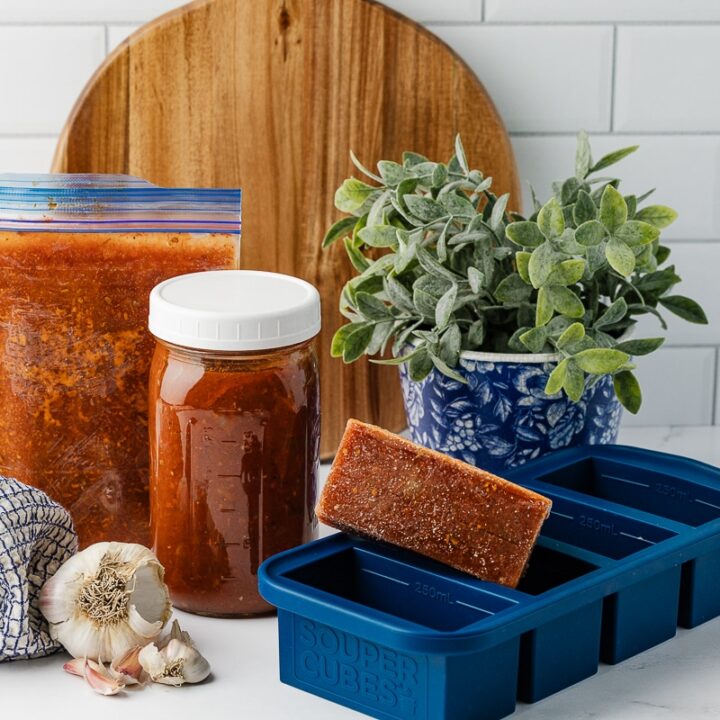 A batch of homemade Everything Tomato Sauce stored in a glass jar, a flat-lay freezer bag, and frozen into Souper Cubes on a white countertop with fresh garlic.