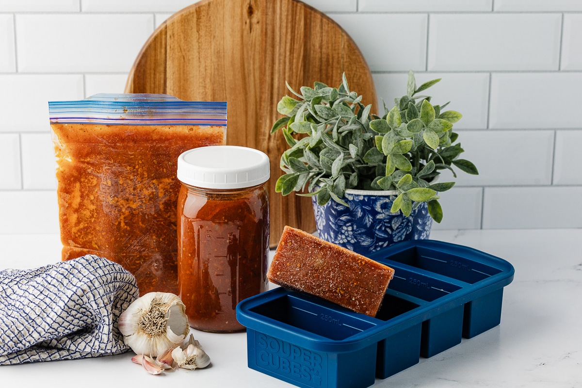 A batch of homemade Everything Tomato Sauce stored in a glass jar, a flat-lay freezer bag, and frozen into Souper Cubes on a white countertop with fresh garlic.