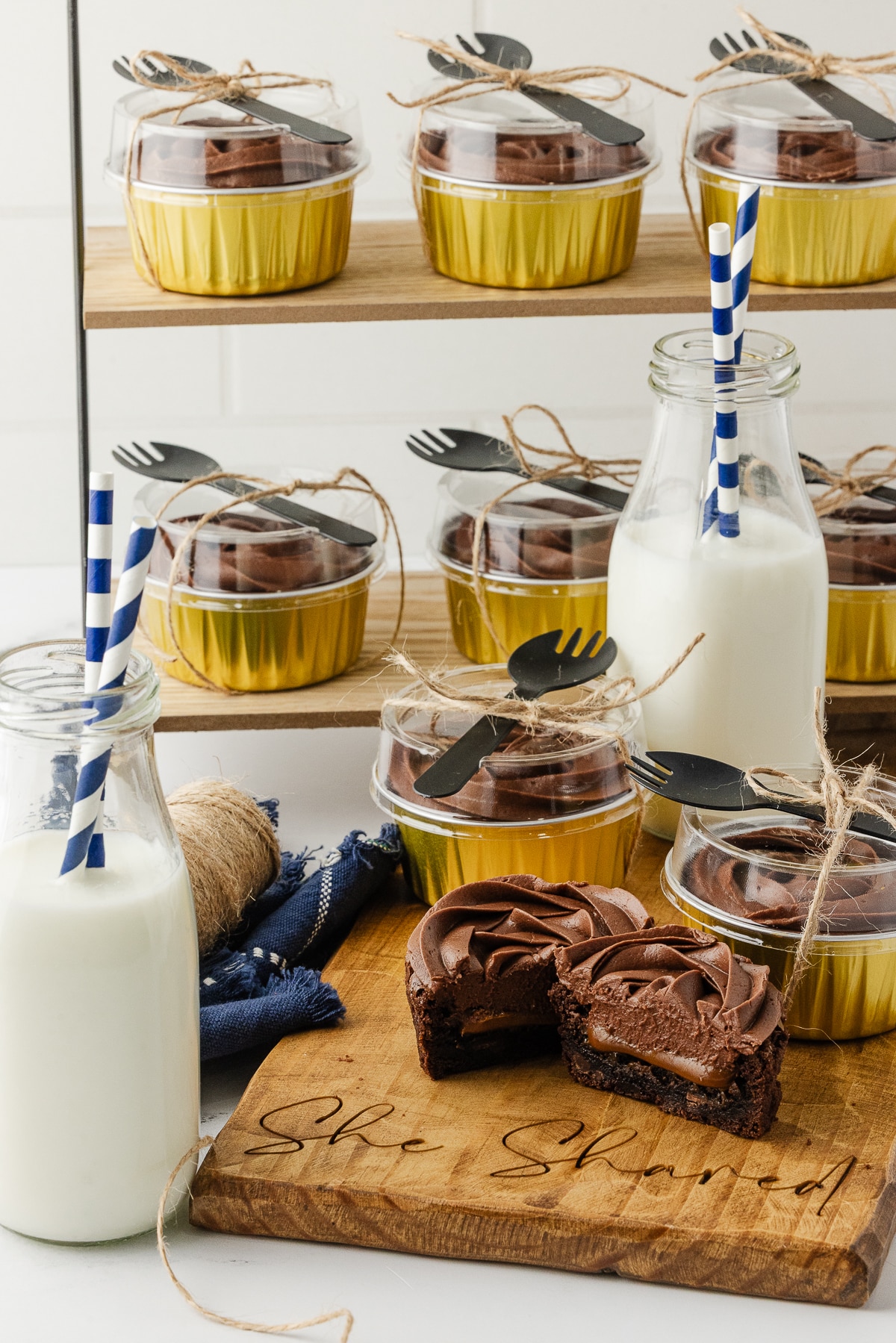 Lifestyle shot of individual gluten-free brownies in gold tins with lids, tied with twine and black sporks, served with bottles of milk on a wooden board.