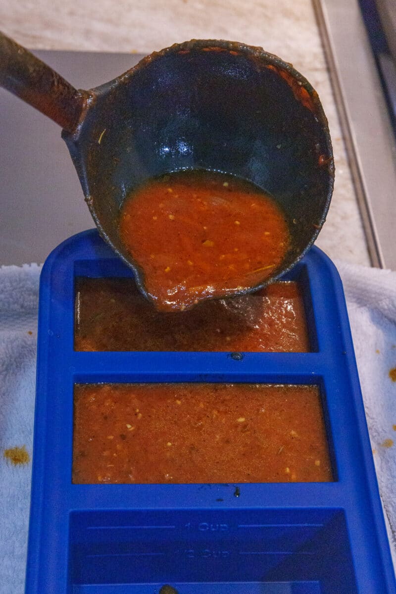 Action shot of a black ladle pouring thick tomato sauce into a silicone freezer tray.