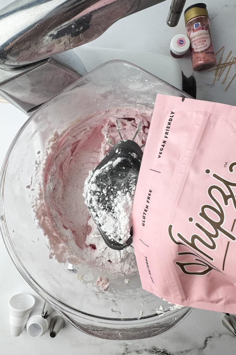 Adding more strawberry Whipzi powdered sugar to the buttercream frosting in a mixer bowl.