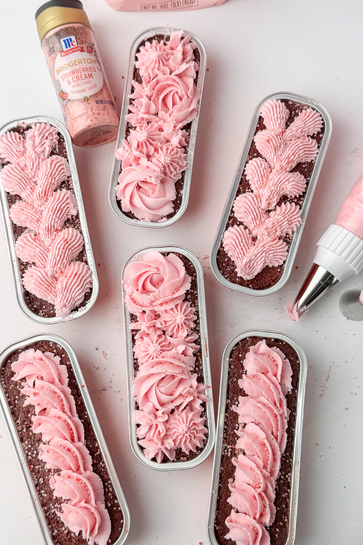 Six chocolate mini loaves with pink strawberry frosting rosettes and braids, styled with a piping bag.