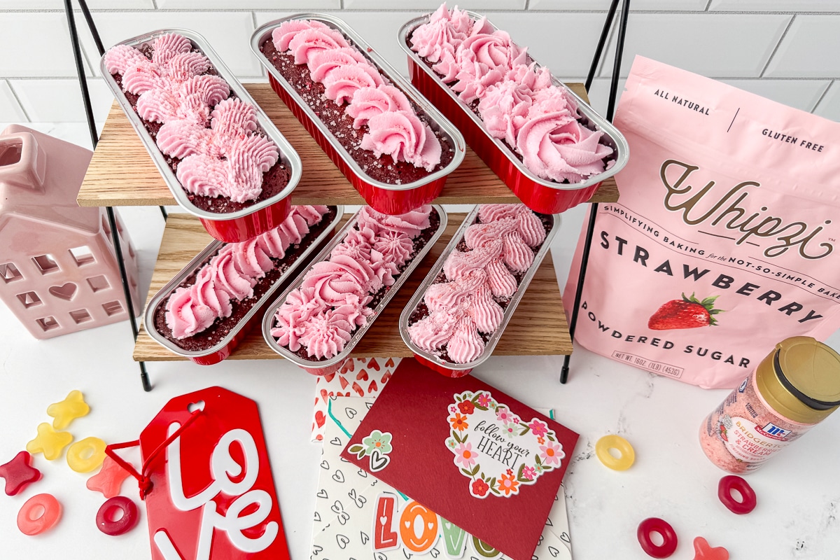 A two-tier wooden stand displaying frosted chocolate mini loaves next to a bag of Whipzi strawberry sugar.