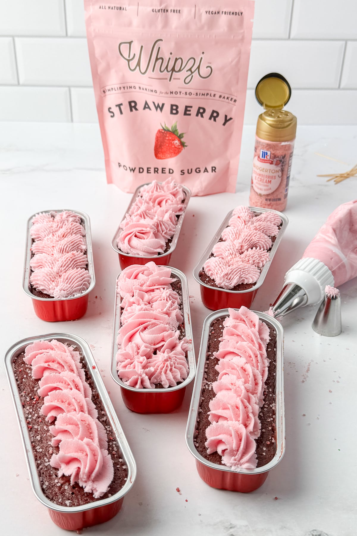 Six chocolate mini loaves with pink frosting arranged in a circle around a bag of Whipzi powdered sugar.