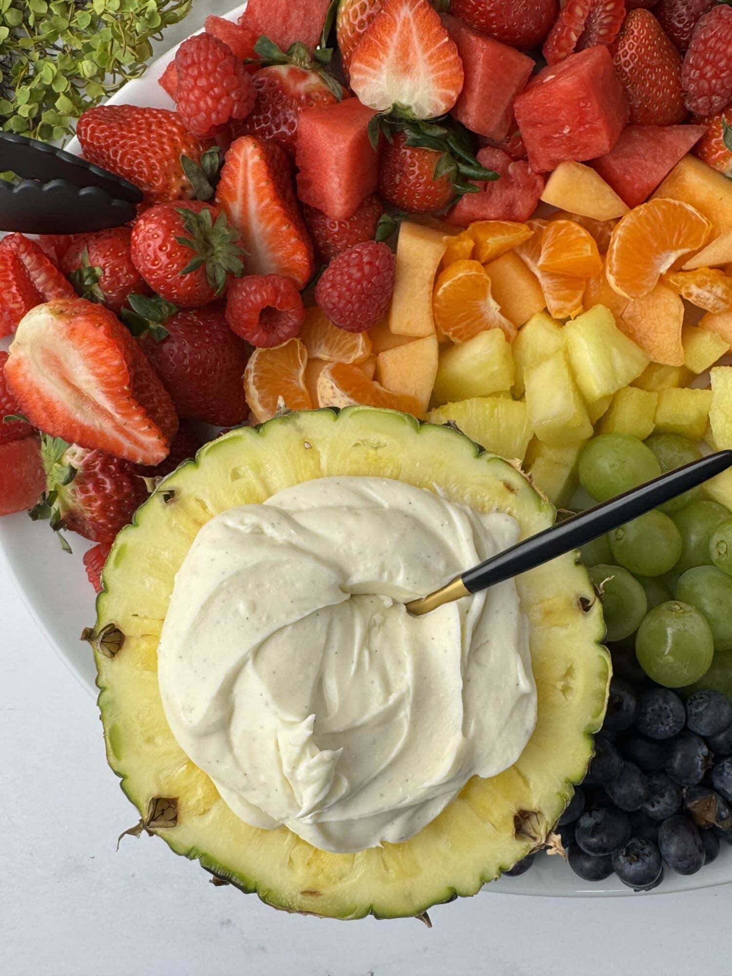 rainbow of fruit surrounding a halved pineapple filled with creamy and tangy fruit dip with a spoon in it