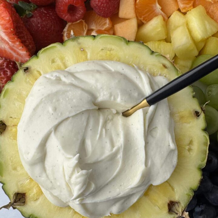 A close-up, top-down view of smooth, velvety cream cheese fruit dip with visible vanilla bean specks, served inside a hollowed-out fresh pineapple and surrounded by strawberries, raspberries, and cantaloupe.
