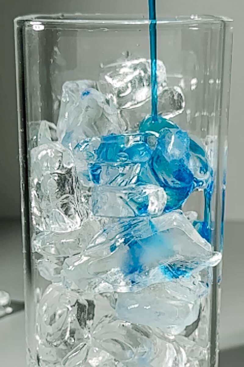 Pouring blue raspberry syrup over ice in a tall glass for a dirty soda recipe base.