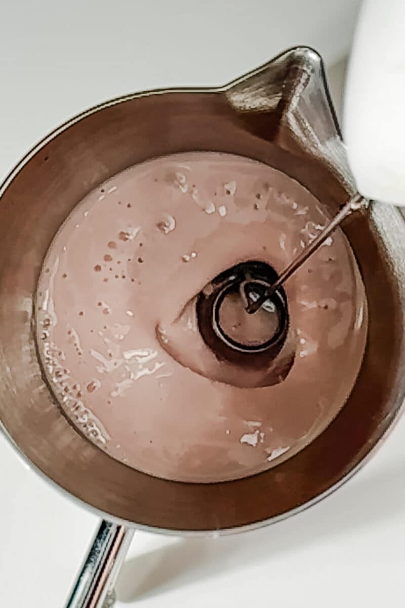 Top-down view of frothing strawberry syrup and heavy cream in a metal pitcher.