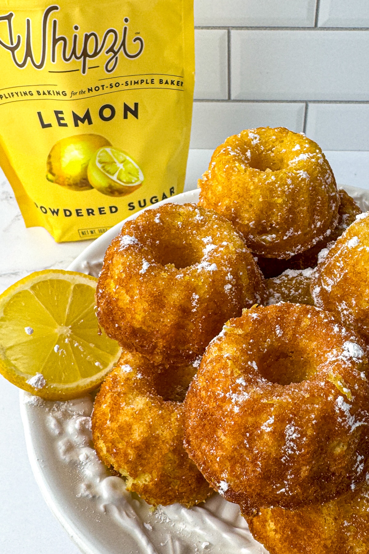 A stack of golden mini lemon drizzle bundt cakes next to a bag of Whipzi lemon powdered sugar and a fresh lemon slice.