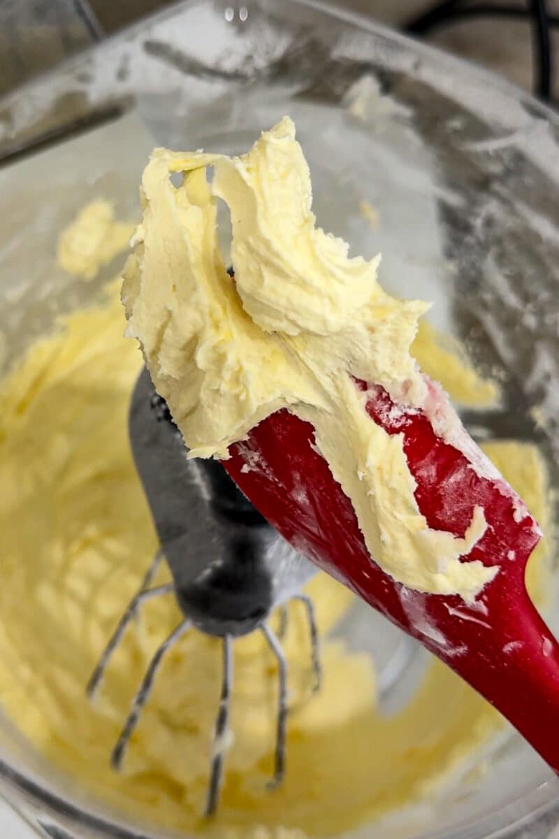 A red rubber spatula holding a creamy, pale yellow mixture of butter and lemon sugar in a stand mixer bowl.