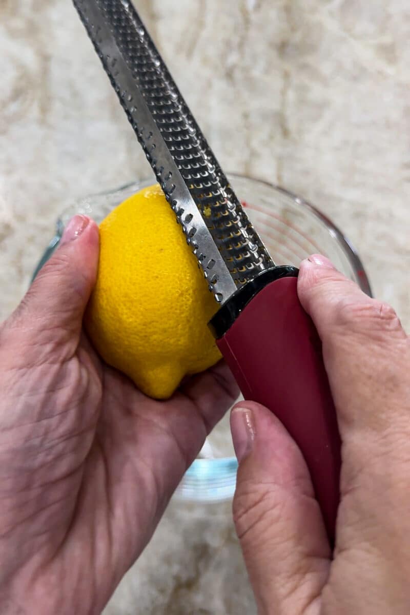 Hands using a zester to grate fresh lemon peel into a bowl for a bright, flavorful mini lemon drizzle cake recipe.