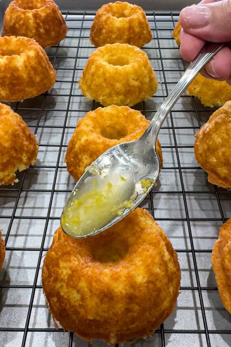 A metal spoon pouring a glossy, sweet lemon drizzle glaze over the top of a freshly baked mini bundt cake.