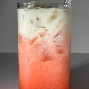 close up of A layered raspberry vanilla lemon-lime dirty soda in a clear glass with ice, showing a bright pink carbonated gradient and a thick, velvety white heavy cream float cascading down from the top.