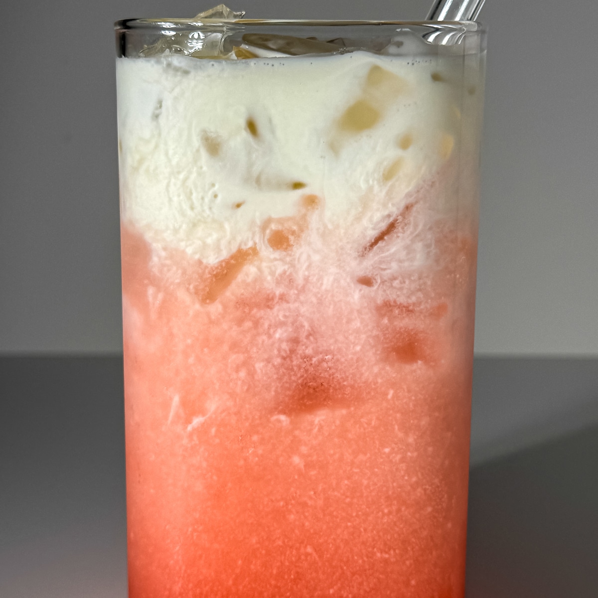 close up of A layered raspberry vanilla lemon-lime dirty soda in a clear glass with ice, showing a bright pink carbonated gradient and a thick, velvety white heavy cream float cascading down from the top.