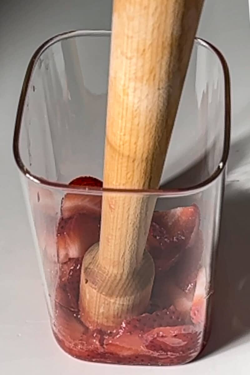 Wooden muddler crushing fresh strawberries in a glass for a homemade dirty soda base.