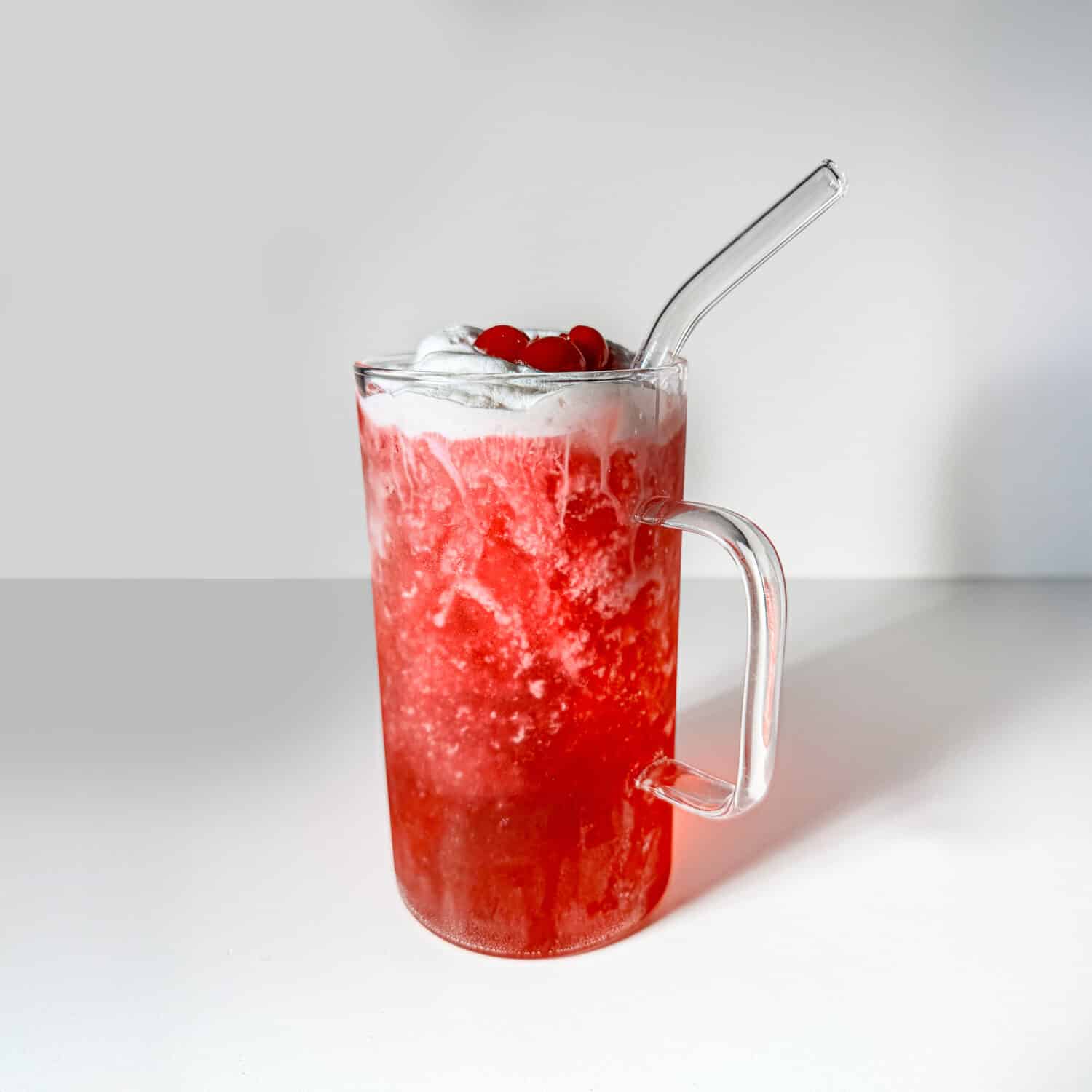 A bright red, bubbly Vanilla Shirley Temple dirty soda mug filled with ice and topped with thick white canned cold foam and maraschino cherries