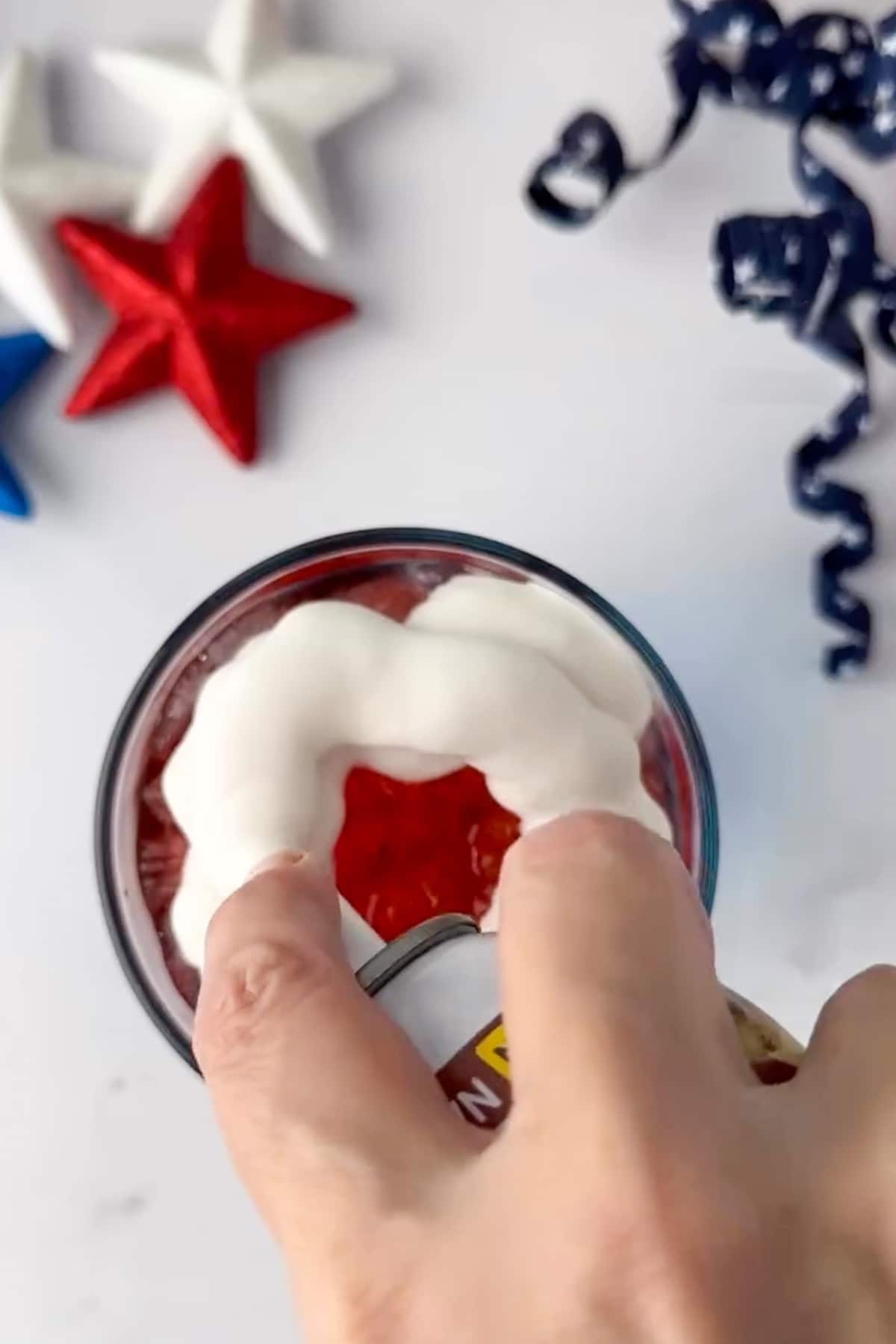 A clean top-down photograph showing a hand spraying sweet canned cold foam in a circular pattern over the red layer of a patriotic mocktail. The crisp white foam contrasts with the rich red strawberry syrup below. In the upper corner, red, white, and blue star decorations rest on the white counter space under natural window lighting.