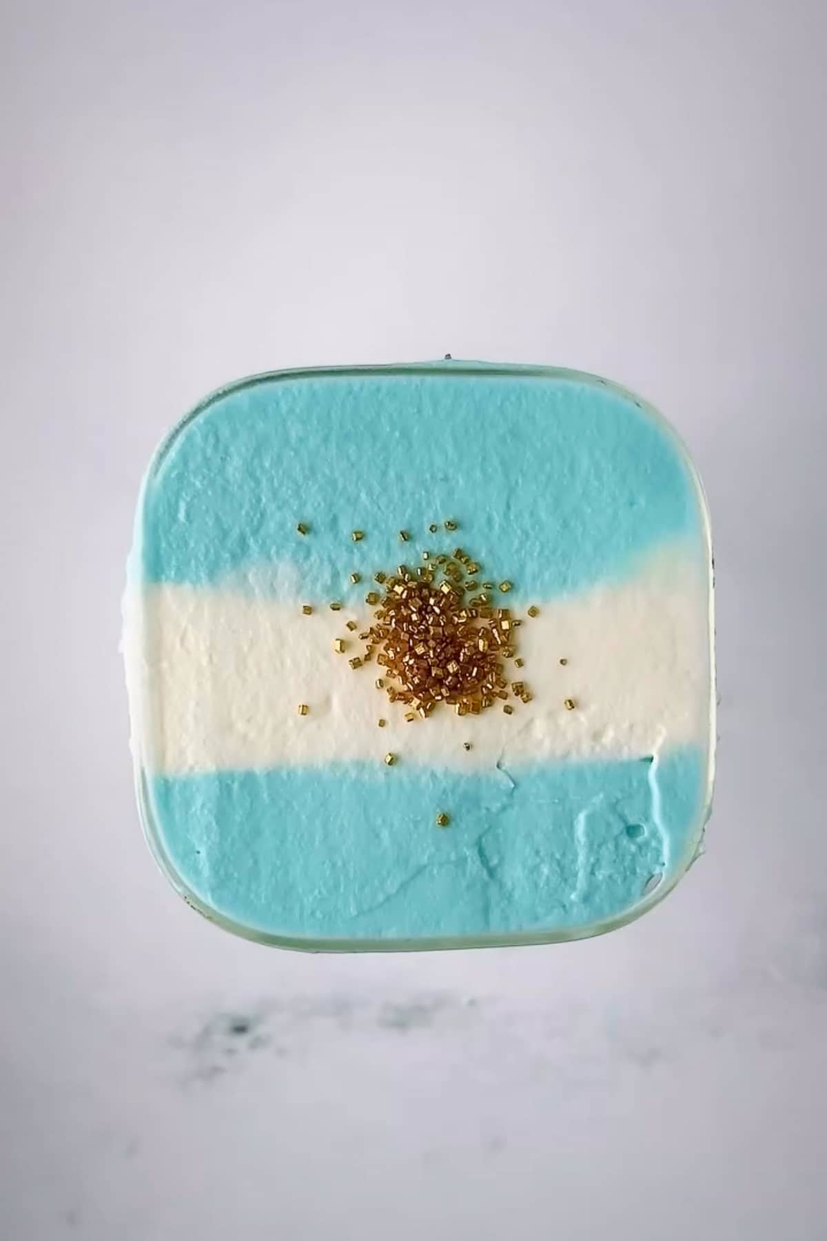 A crisp, macro top-down photograph of a square glass filled with an Argentina flag dirty soda. The surface is layered with thick, velvety homemade cold foam styled into three distinct horizontal stripes—sky blue on the top and bottom, and snowy white through the center. A neat mound of metallic gold Wilton sugar crystals sits perfectly in the middle of the white stripe to represent the flag's sun emblem.