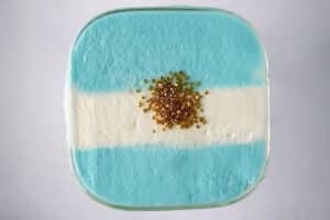 A close-up, high-realism shot looking directly down at the square rim of a blue raspberry dirty soda. The top features a stable, hand-frothed heavy cream topping arranged in the horizontal pattern of the Argentine flag. Shimmering gold sugar crystals are clustered right in the center over the white foam stripe, catching natural, directional window light that casts soft textures across the cream.