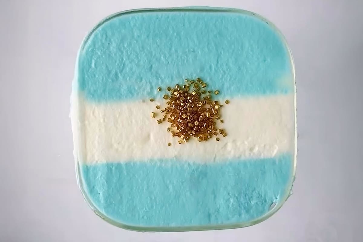 A close-up, high-realism shot looking directly down at the square rim of a blue raspberry dirty soda. The top features a stable, hand-frothed heavy cream topping arranged in the horizontal pattern of the Argentine flag. Shimmering gold sugar crystals are clustered right in the center over the white foam stripe, catching natural, directional window light that casts soft textures across the cream.