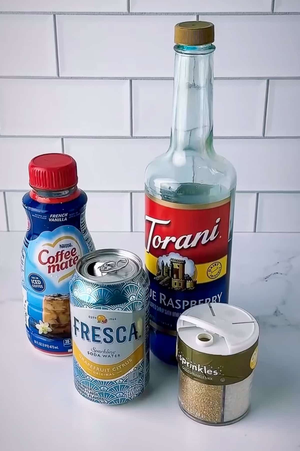 A bright counter lineup of the exact tools and ingredients used for the Day 5 dirty soda recipe. A silver can of zero-sugar Grapefruit Citrus Fresca sits in the foreground next to a bottle of gold Wilton sprinkles. Behind them is a large bottle of Torani Blue Raspberry syrup and a container of French Vanilla creamer, set against a classic white subway tile kitchen backsplash.